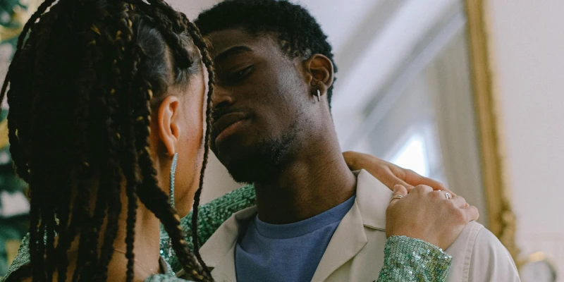 Couple Embracing by the Mirror A couple is hugging in front of a mirror. One person is wearing a sparkly green dress with braided hair, while the other wears a light-colored jacket over a blue shirt. A festive Christmas tree is partially visible in the background.